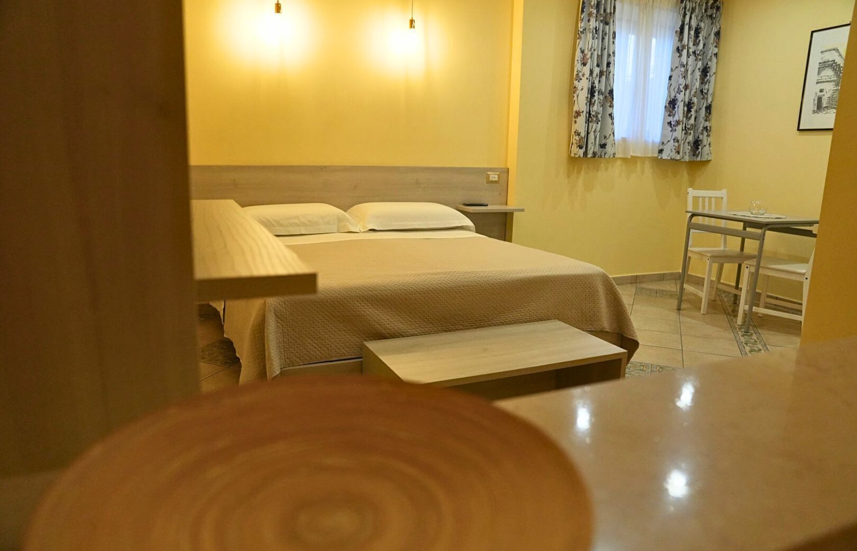 Una camera d'albergo con un letto, un comodino, una lampada, una sedia e un tavolino con una seconda sedia.