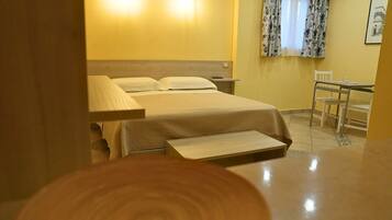 Una camera d'albergo con un letto, un comodino, una lampada, una sedia e un tavolino con una seconda sedia.