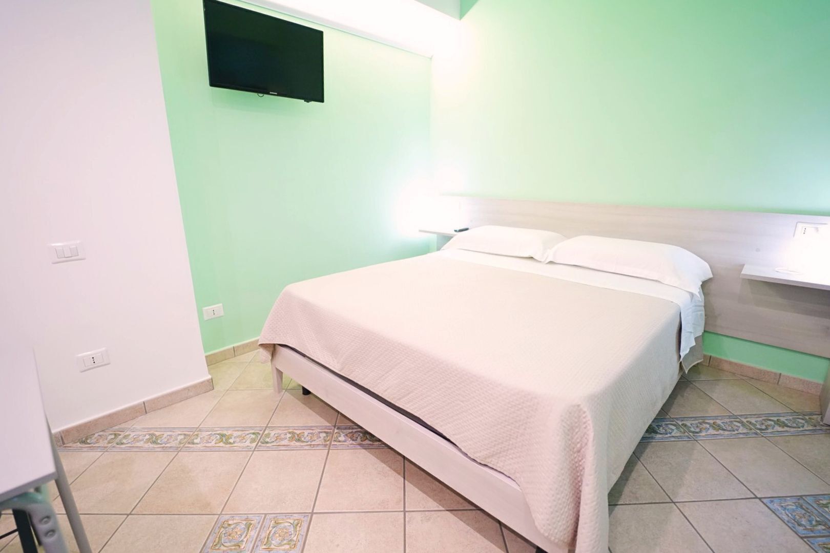 A bedroom with a bed, a bedside table, a TV mounted on the wall, and a tiled floor.