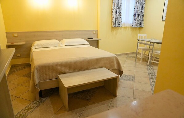 Una habitación de hotel con cama, mesitas de noche, un banco, una pequeña mesa y una silla.