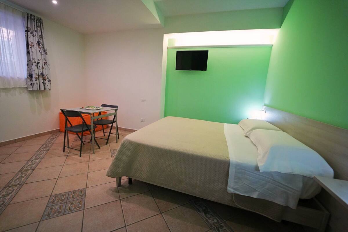 A room with a bed, a desk with a chair, a television on the wall, and a patterned floor.