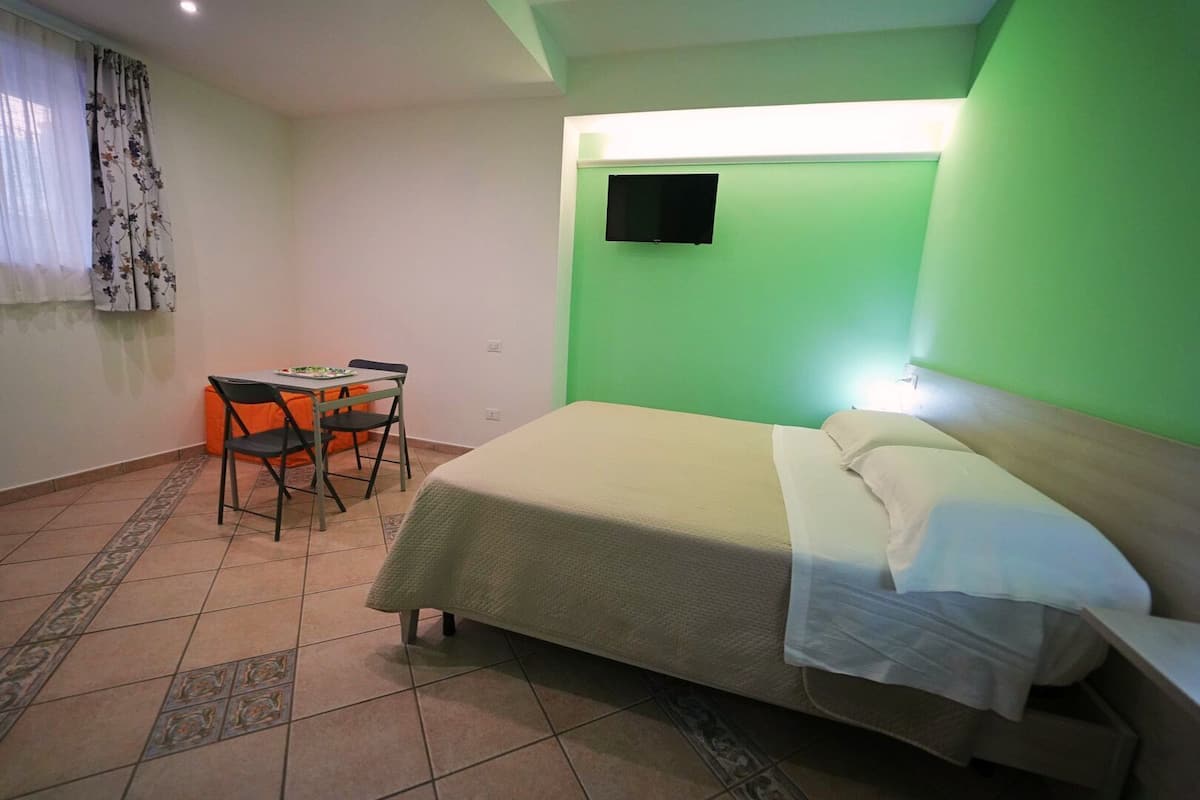 A room with a bed, a desk with a chair, a flat-screen TV mounted on the wall, and a patterned rug on the floor.