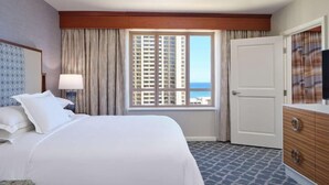 A hotel room with a bed, a dresser, a lamp, a window with a view of buildings, and a door. - Cozy 1-bedroom apartment in marvelous Honolulu with AC and gym Grand Waikikian (Honolulu)