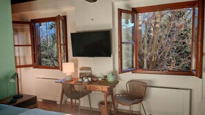 Une chambre comprenant un lit, un bureau avec des chaises, une télévision et des fenêtres donnant sur des arbres.