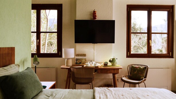 Une chambre à coucher comprenant un lit, un bureau avec une chaise, un téléviseur fixé au mur et des fenêtres donnant sur des arbres.