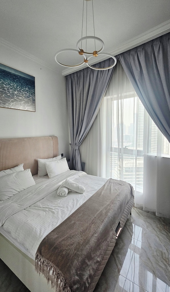 A modern hotel room with a large bed, a headboard, a painting on the wall, and a view of the city through the window. - Upgraded, sparkling clean 1 BR apt. w/balcony. Full Marina view. 5 min to beach (Dubai)