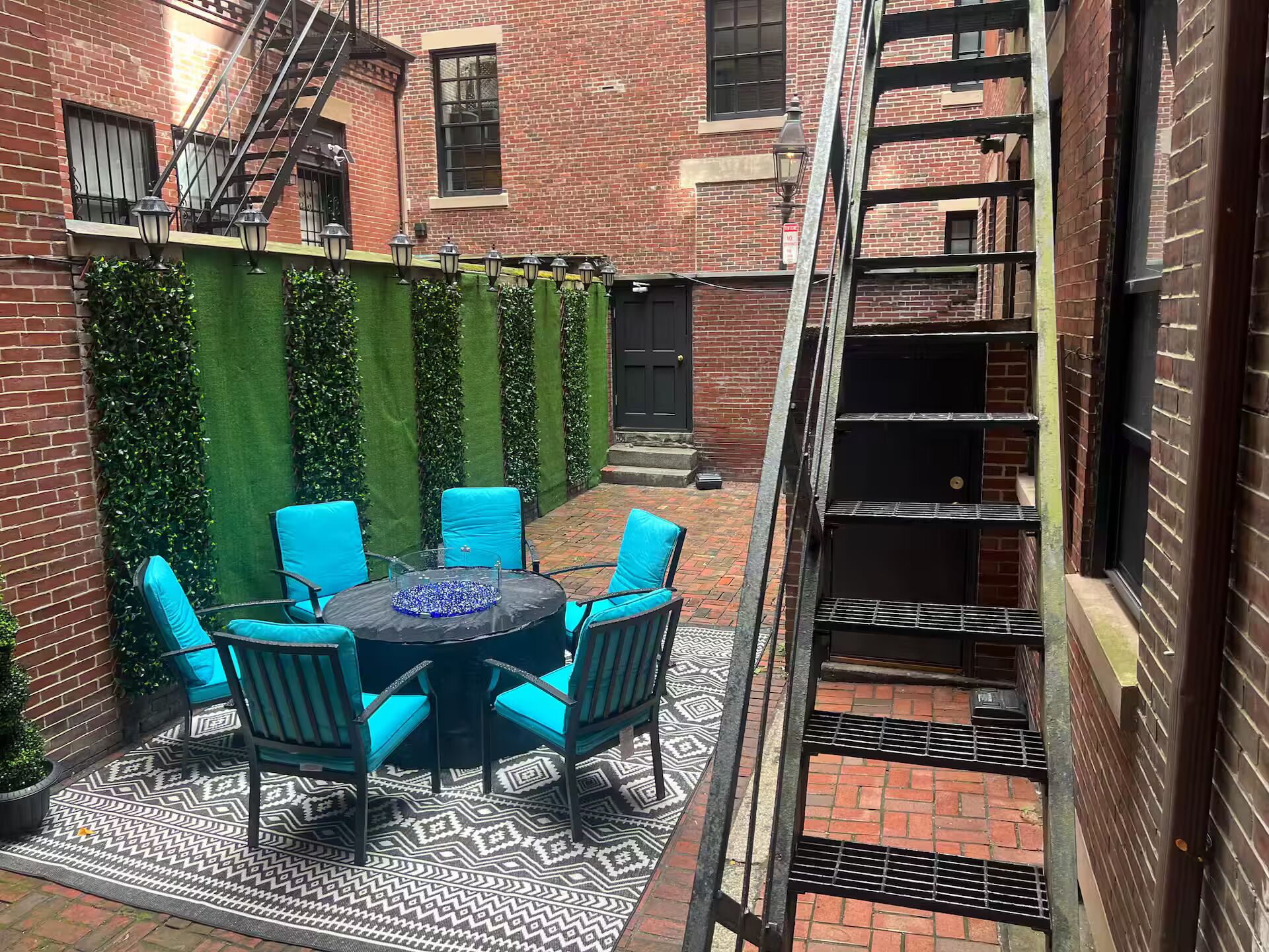 A courtyard with a round table and chairs, a brick building, a green wall, and a metal staircase.
