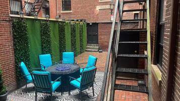 A courtyard with a round table and chairs, a brick building, a green wall, and a metal staircase.