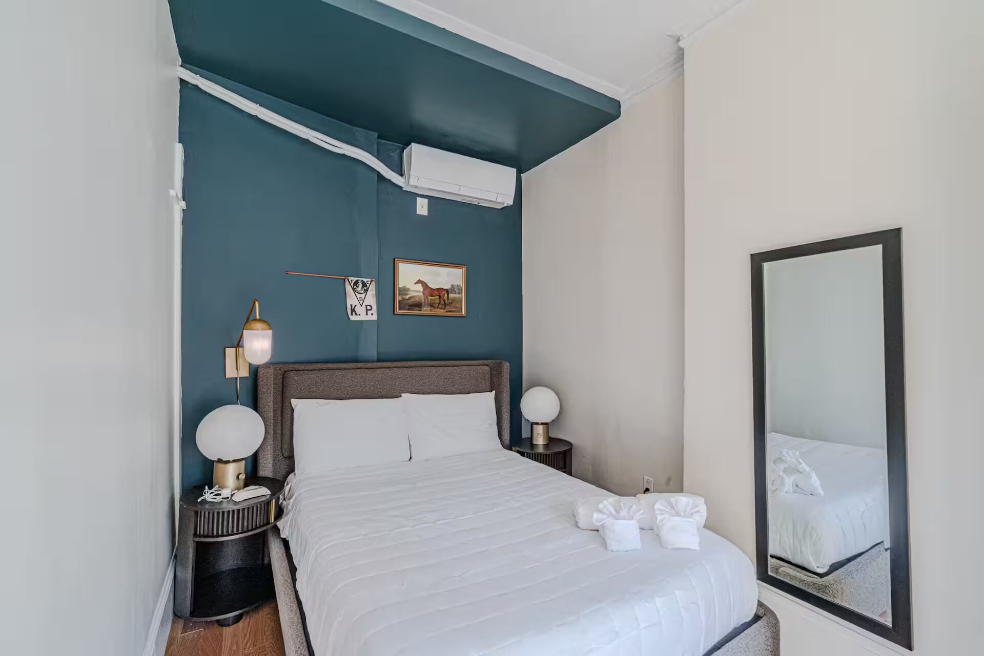 A hotel room with a bed, bedside tables, a mirror, and a wall-mounted air conditioning unit.