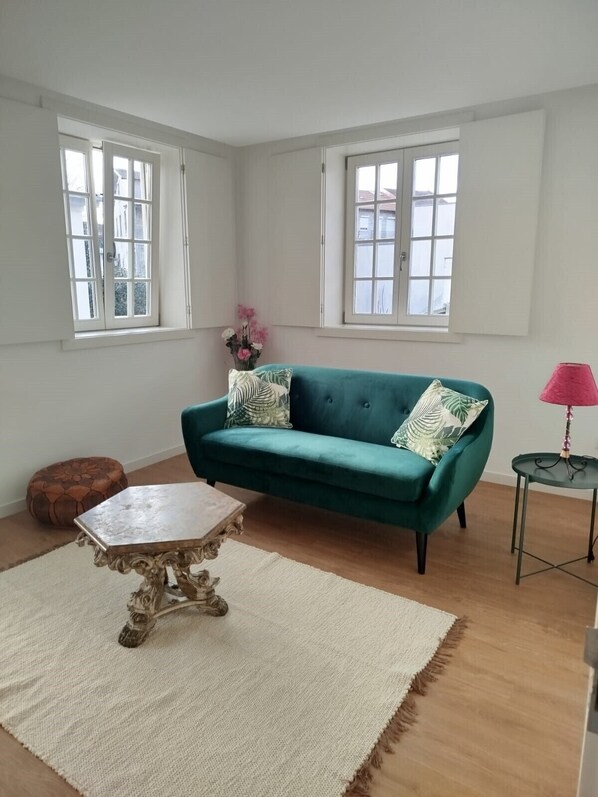 A room with a green sofa, a small round table, a decorative table with ornate legs, a pink lamp, and a vase with flowers.