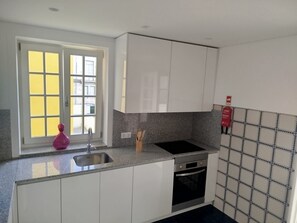 A kitchen with white cabinets, a stainless steel sink, a black oven, a window with yellow blinds, and a tiled backsplash.