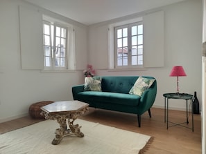 A room with a green sofa, a small round table, a white rug, and two windows with white frames. - 4 - The Floor House 4 Bedrooms Center (Guimarães)
