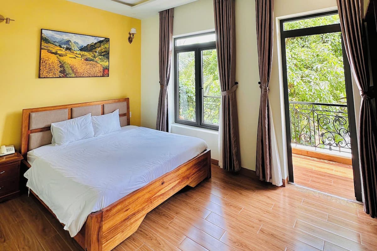 A bedroom with a wooden bed, a painting on the wall, a window with curtains, and a balcony with a railing.