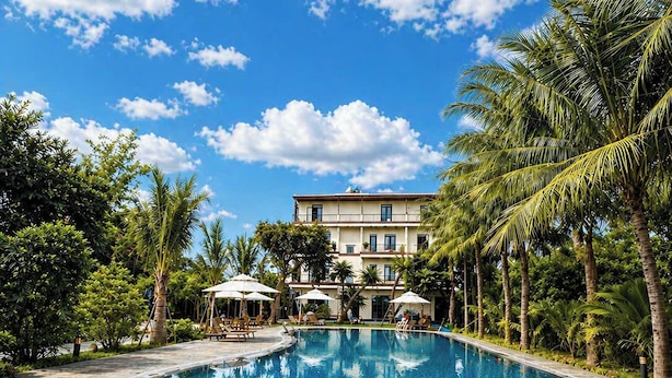 Una piscina con borde de azulejos, rodeada de palmeras y un edificio con balcón.
