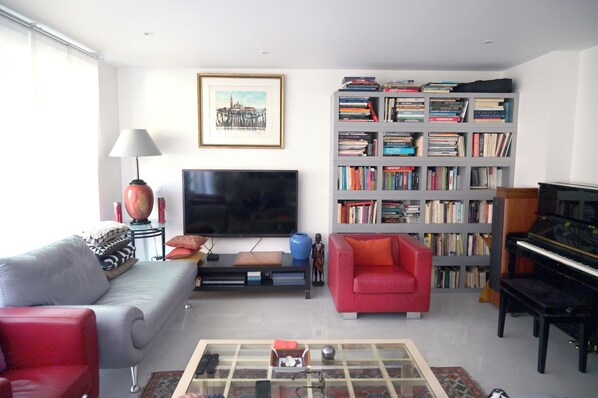 A living room with a sofa, a red armchair, a bookshelf, a television, a piano, and a coffee table.