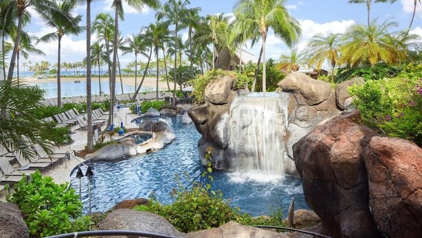 En pool på ett semesterhotell med ett vattenfall, palmer och utsikt över stranden.