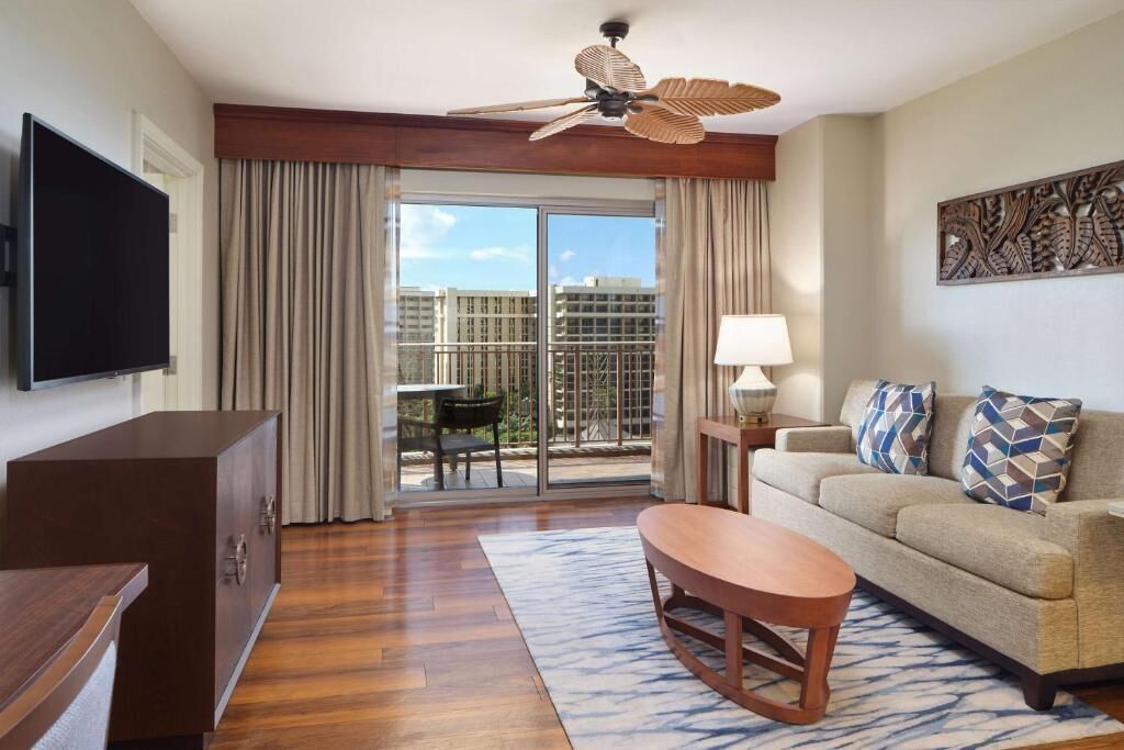 A hotel room with a flat-screen TV, a ceiling fan, a sofa with patterned cushions, a wooden coffee table, and a view of a cityscape through the balcony doors.