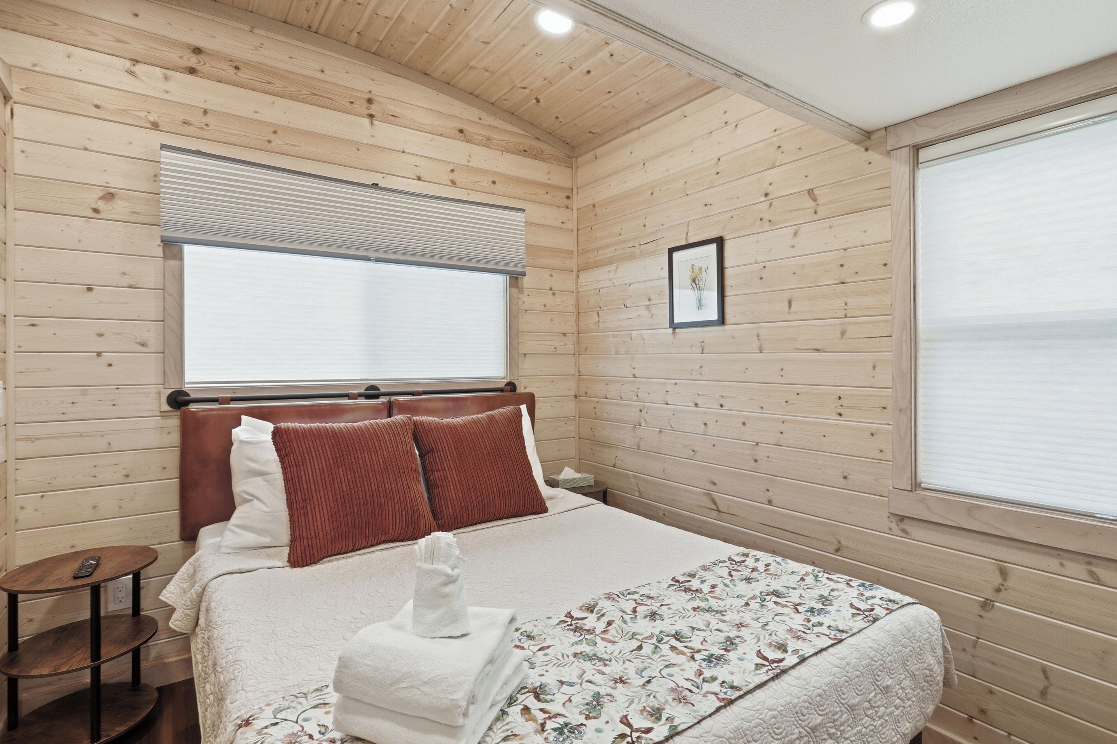 A wooden-paneled room with a bed, a bedside table, a window with blinds, and a framed picture on the wall.