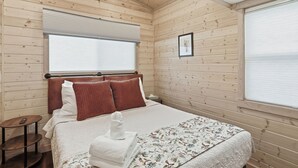 Habitación con paneles de madera, una cama, una mesita de noche, una ventana con persianas y un cuadro enmarcado en la pared.