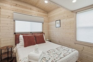 Habitación con paneles de madera, una cama, una mesita de noche, una ventana con persianas y un cuadro enmarcado en la pared.