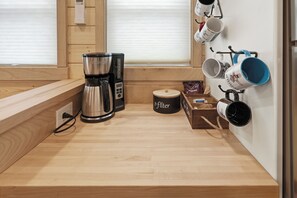 A compact kitchen with a coffee maker, a kettle, and a coffee grinder. There are mugs hanging on the wall and a small wooden box with a label.