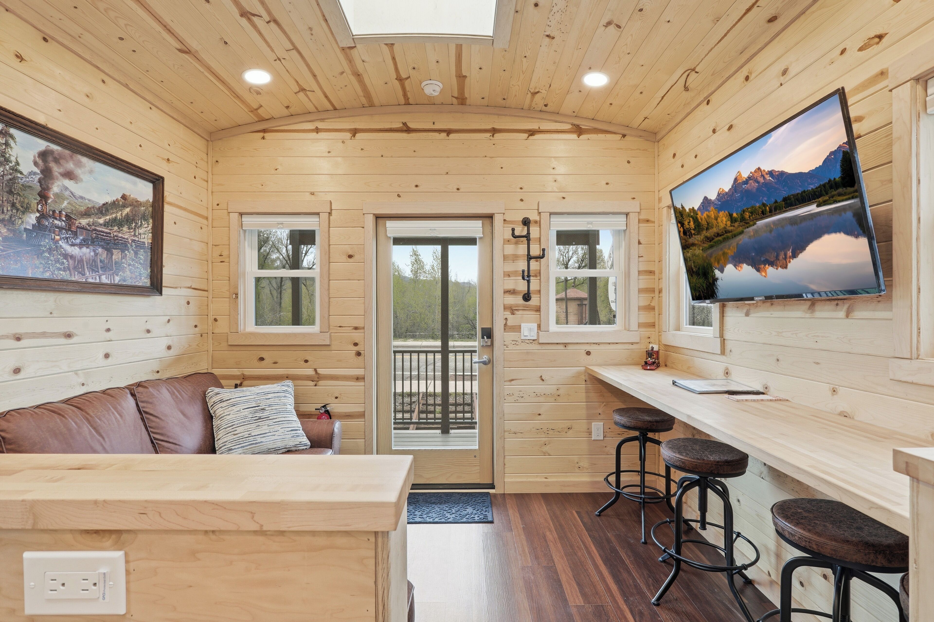 A cabin interior with a wooden ceiling, a couch with pillows, a bar area with stools, and a television on the wall.