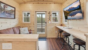 Interior de una cabaña con techo de madera, un sofá con almohadones, una zona de bar con taburetes y un televisor en la pared.
