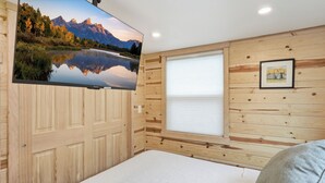 Un dormitorio con cama, ventana con persianas, televisor montado mostrando un paisaje de montaña y un cuadro en la pared.