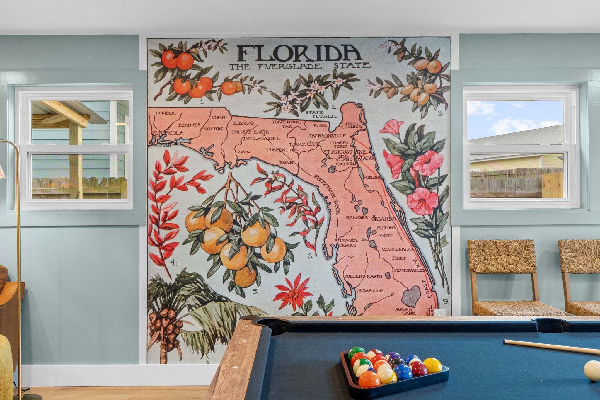 A pool table with a Florida-themed map mural, a window with a view, and two wicker chairs.