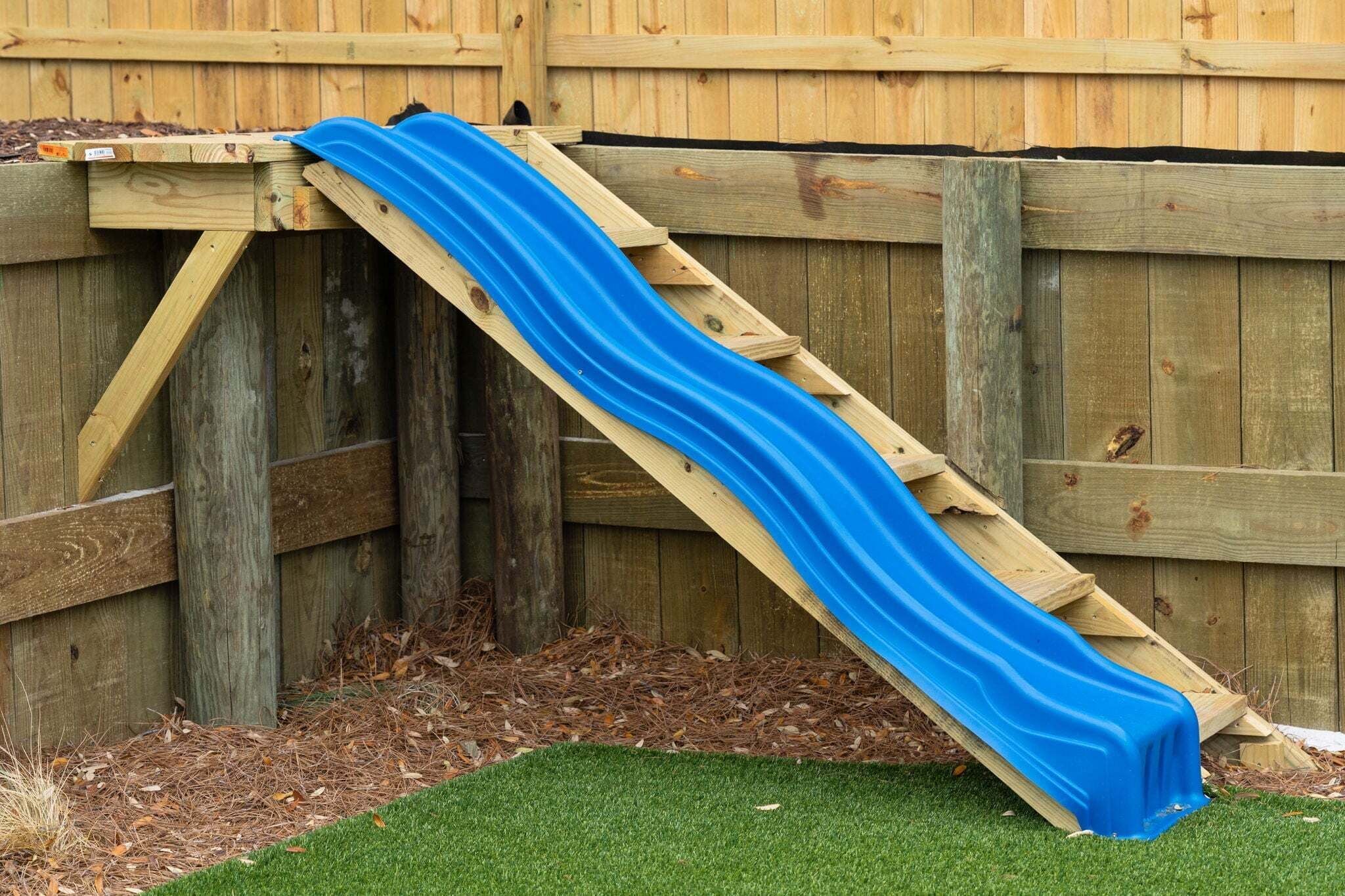 A wooden slide structure with a blue slide against a wooden fence.