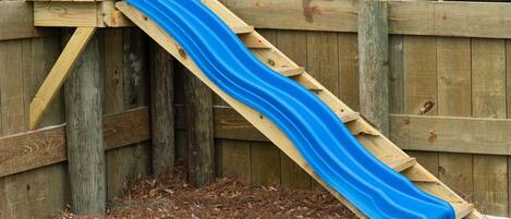 A wooden slide structure with a blue slide against a wooden fence.