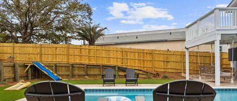 Un patio trasero con piscina, dos sillas Adirondack, un tobogán y una valla de madera.