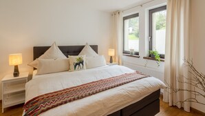 Ein ordentlich bezogenes Bett mit einem gemusterten Überwurf, zwei Nachttischlampen und einem Fenster mit Vorhängen.