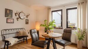 Ein gemütliches Wohnzimmer mit einem runden Holztisch, zwei Ledersesseln, einer schwarzen Metallbank, einer Stehlampe und einem Fenster mit Vorhängen.