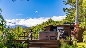 Une terrasse en bois avec des marches menant à un bâtiment, entourée d’une végétation luxuriante et sous un ciel bleu dégagé.