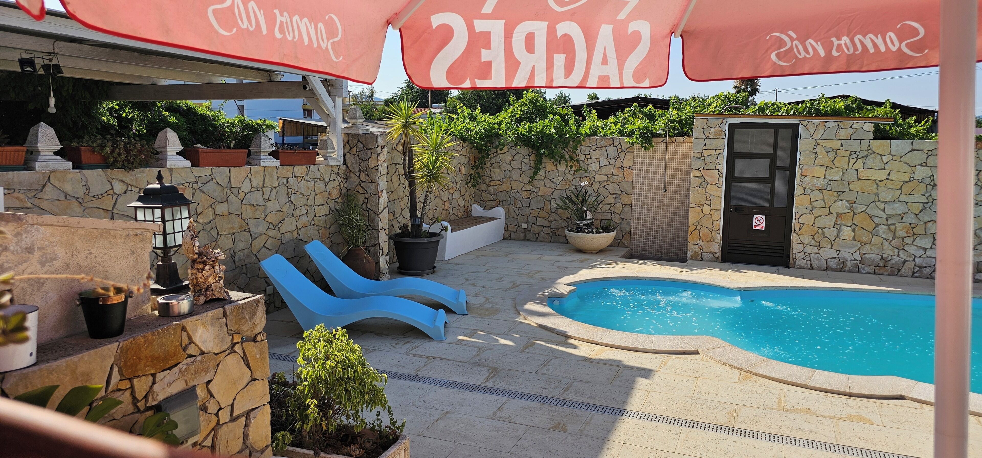 Zona de piscina con pared de piedra, una silla de descanso azul, una planta en maceta y un paraguas rojo.