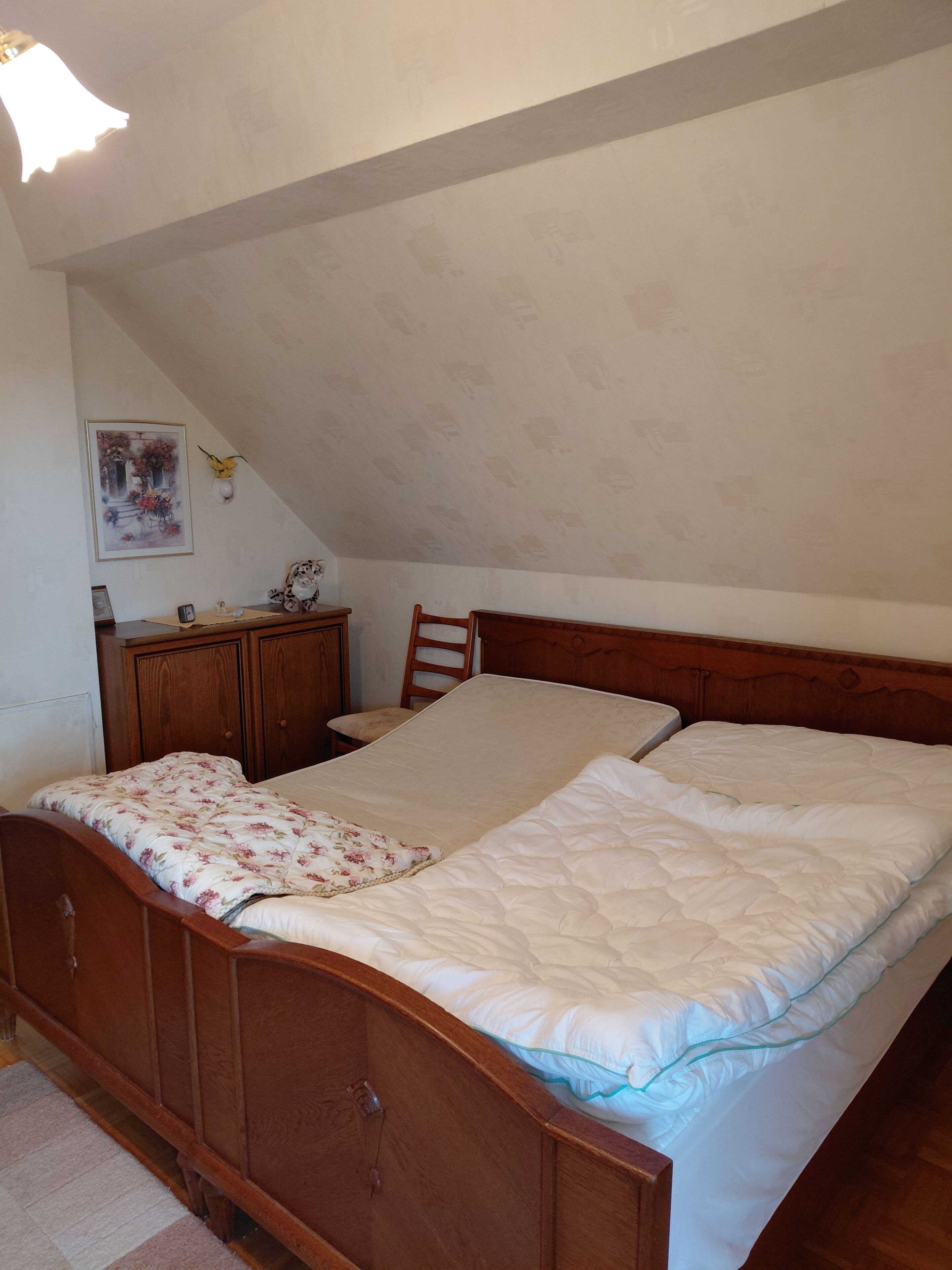 A bedroom with a wooden bed, a nightstand, a chair, a wall-mounted lamp, and a framed picture.