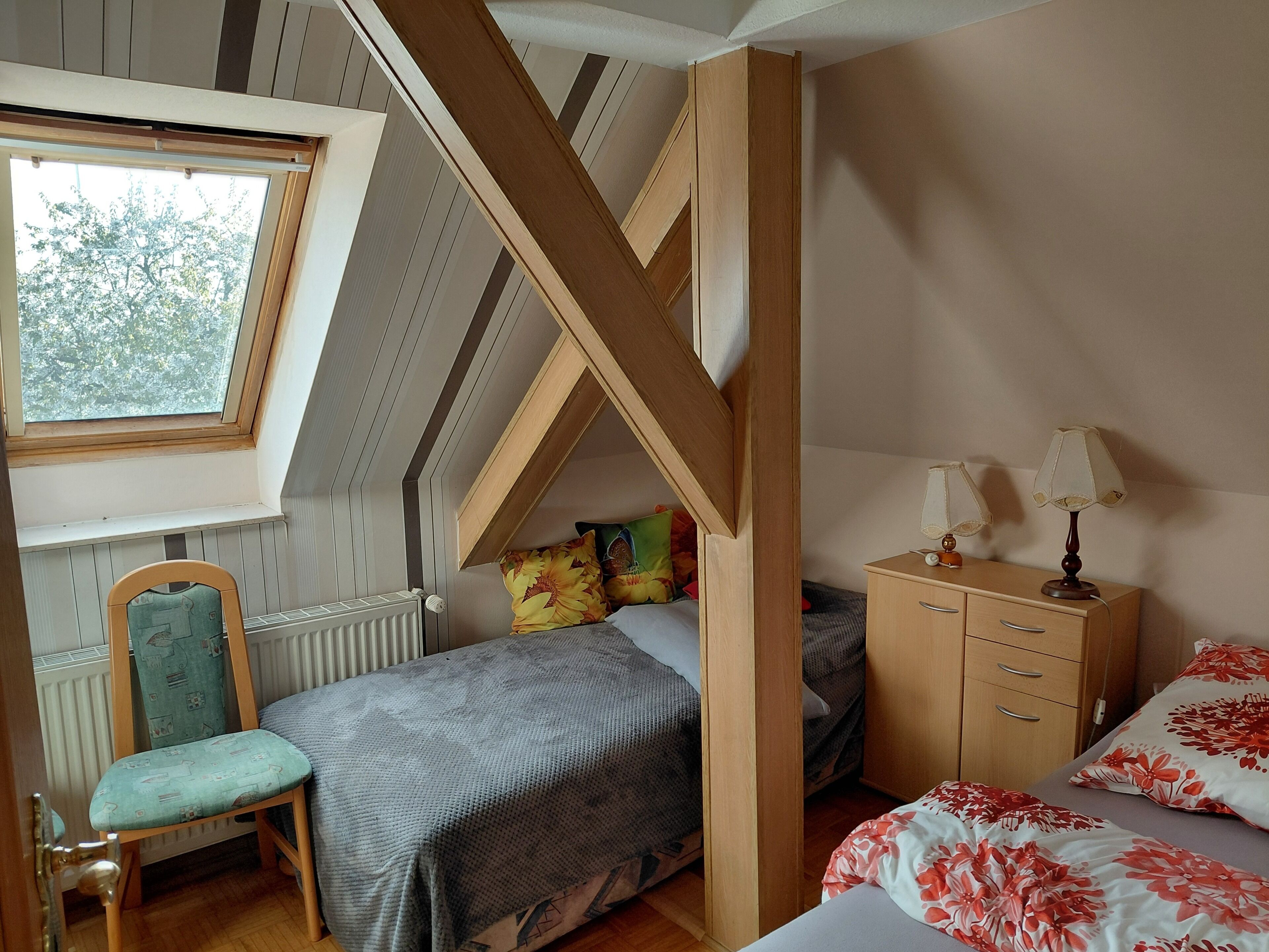 Un dormitorio con techo inclinado, una ventana, una cama con ropa de cama floral, una silla, una mesita de noche con lámparas y un armario de madera.