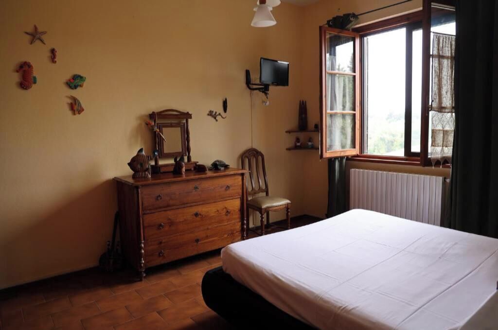 A bedroom with a wooden dresser, a chair, a bed, a television, and a window with curtains.
