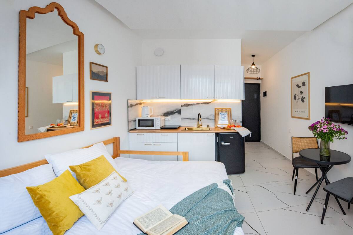 A modern hotel room with a bed, bedside table, a large mirror, a small kitchenette, a dining area with a table and chairs, and a wall-mounted TV.