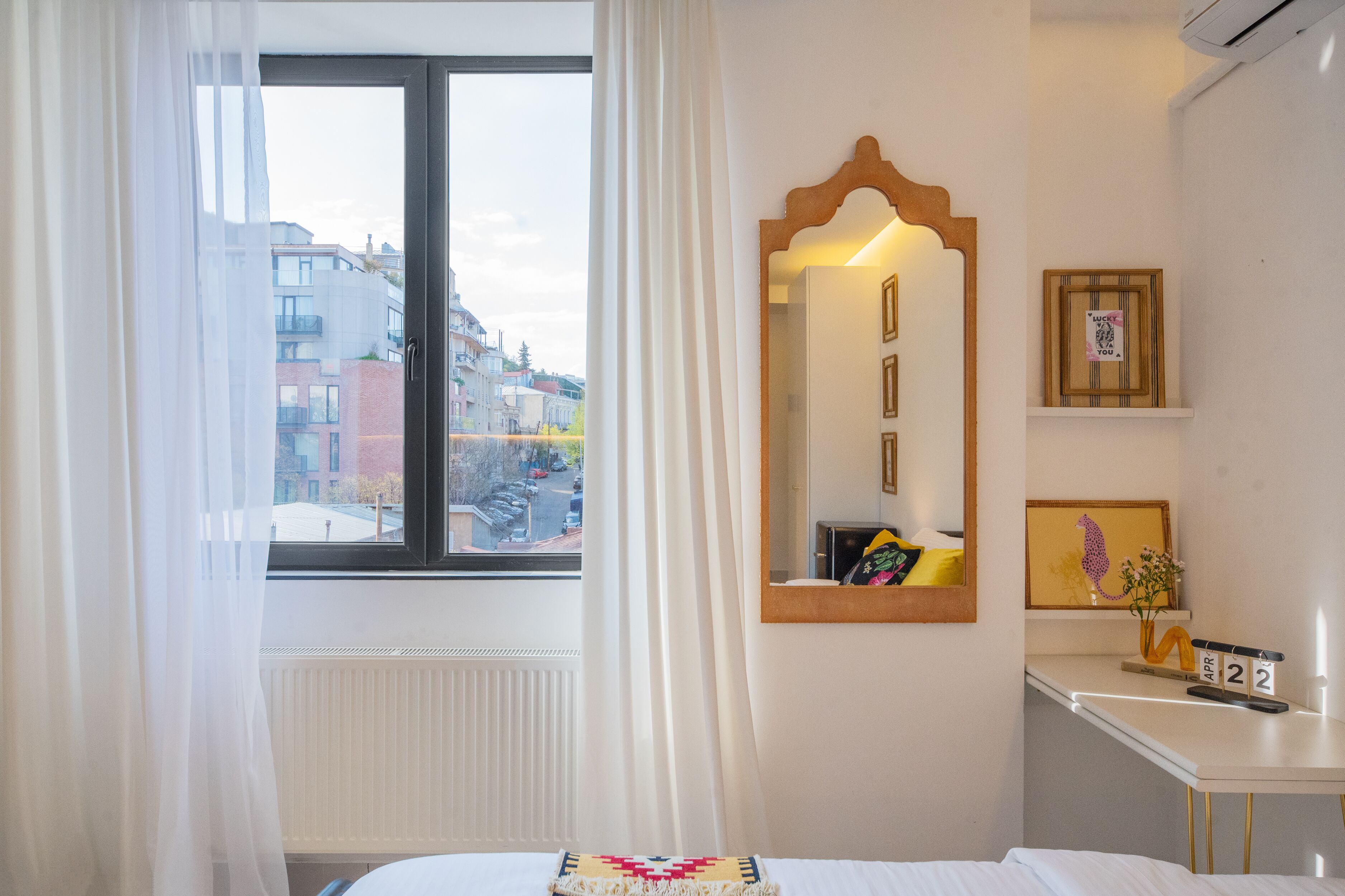 A hotel room with a bed, a window with curtains, a mirror, a desk with a calendar, and a radiator.