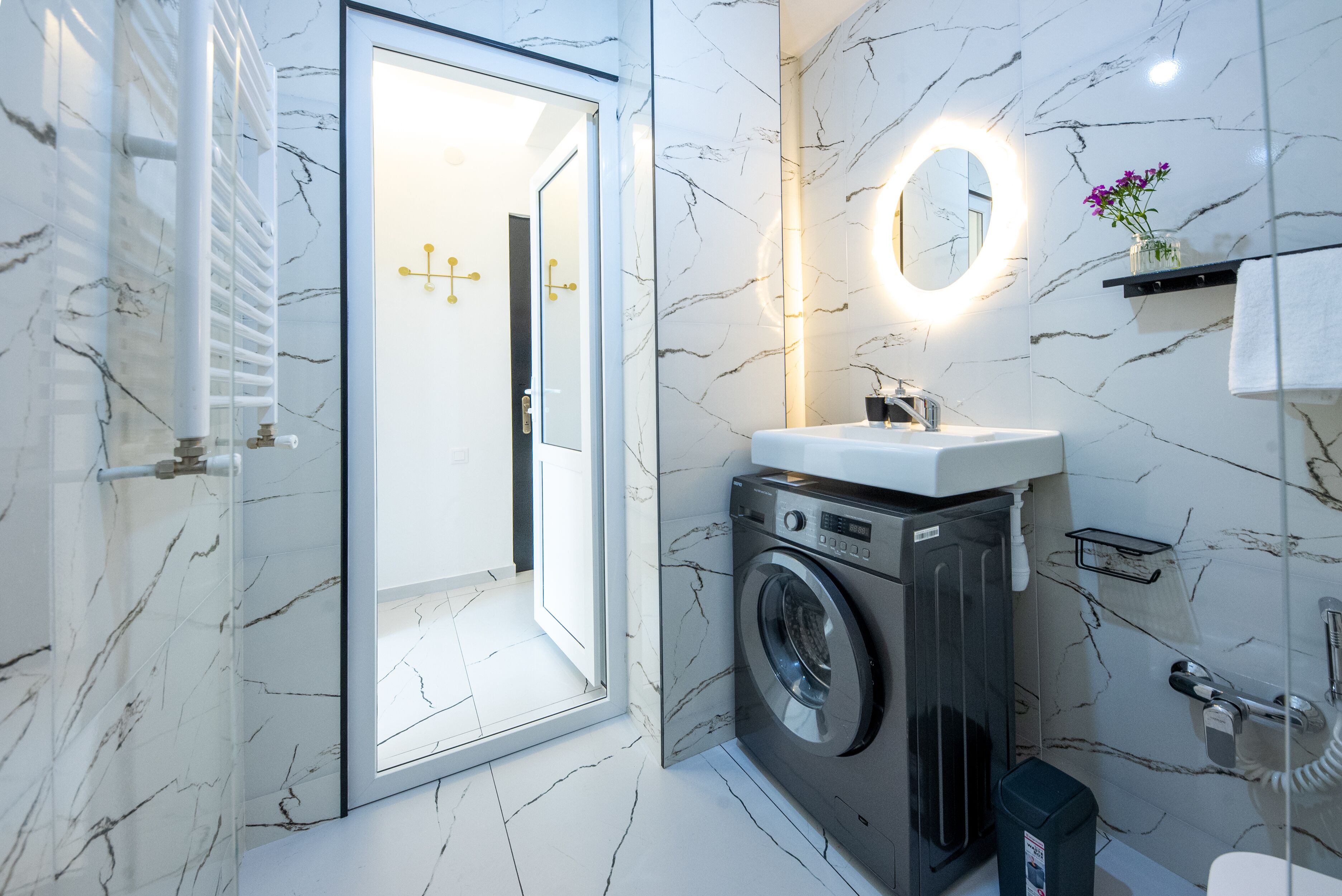 A modern bathroom with a marble wall, a washing machine, a sink, a mirror, and a shower area.