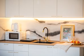 Une cuisine avec des armoires blanches, un pare-feu en marbre, un évier avec un robinet doré, un four à micro-ondes, une machine à café et un tableau encadré.