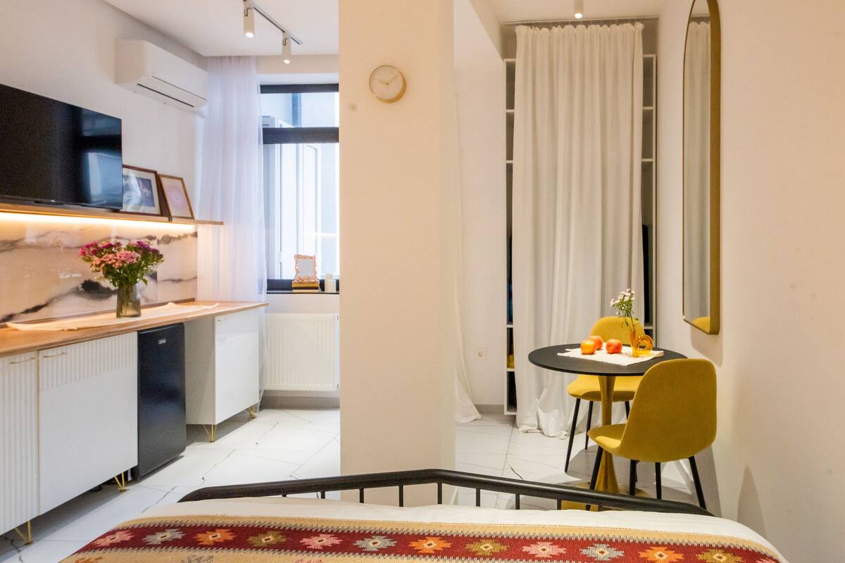 A modern hotel room with a bed, a desk with a computer, a chair, a small table with fruit, and a window with curtains.