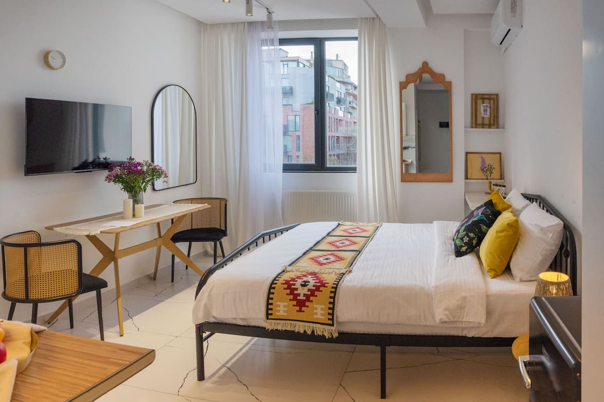 A hotel room with a bed, a desk, a chair, a TV, and a window with a view of buildings.