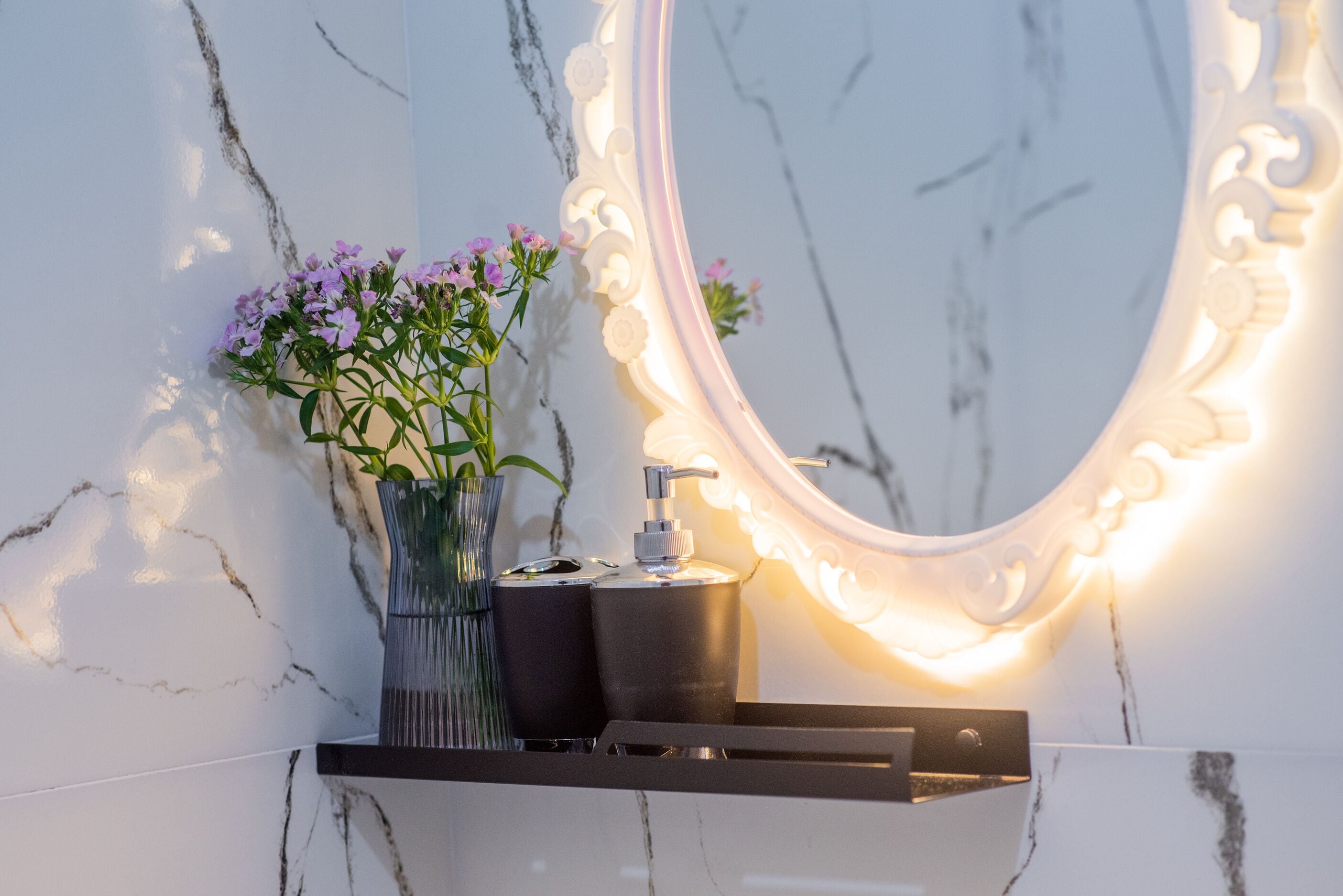 Une salle de bain avec un mur en marbre, un grand miroir, un lavabo doté d’un distributeur de savon noir et un vase contenant des fleurs violettes.