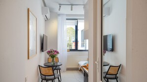 Habitación compacta con cama, una mesita con sillas, televisor montado en la pared y ventana con vistas al exterior.