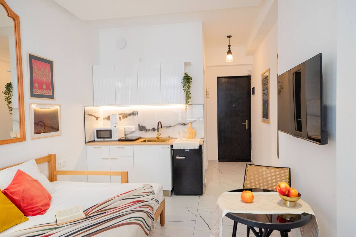 A compact hotel room with a bed, bedside table, a small kitchenette, a dining area with a table and chairs, and a TV mounted on the wall.