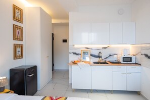 Une chambre d’hôtel compacte comprenant un coin cuisine, un petit réfrigérateur, un lit et une table de chevet avec une lampe.