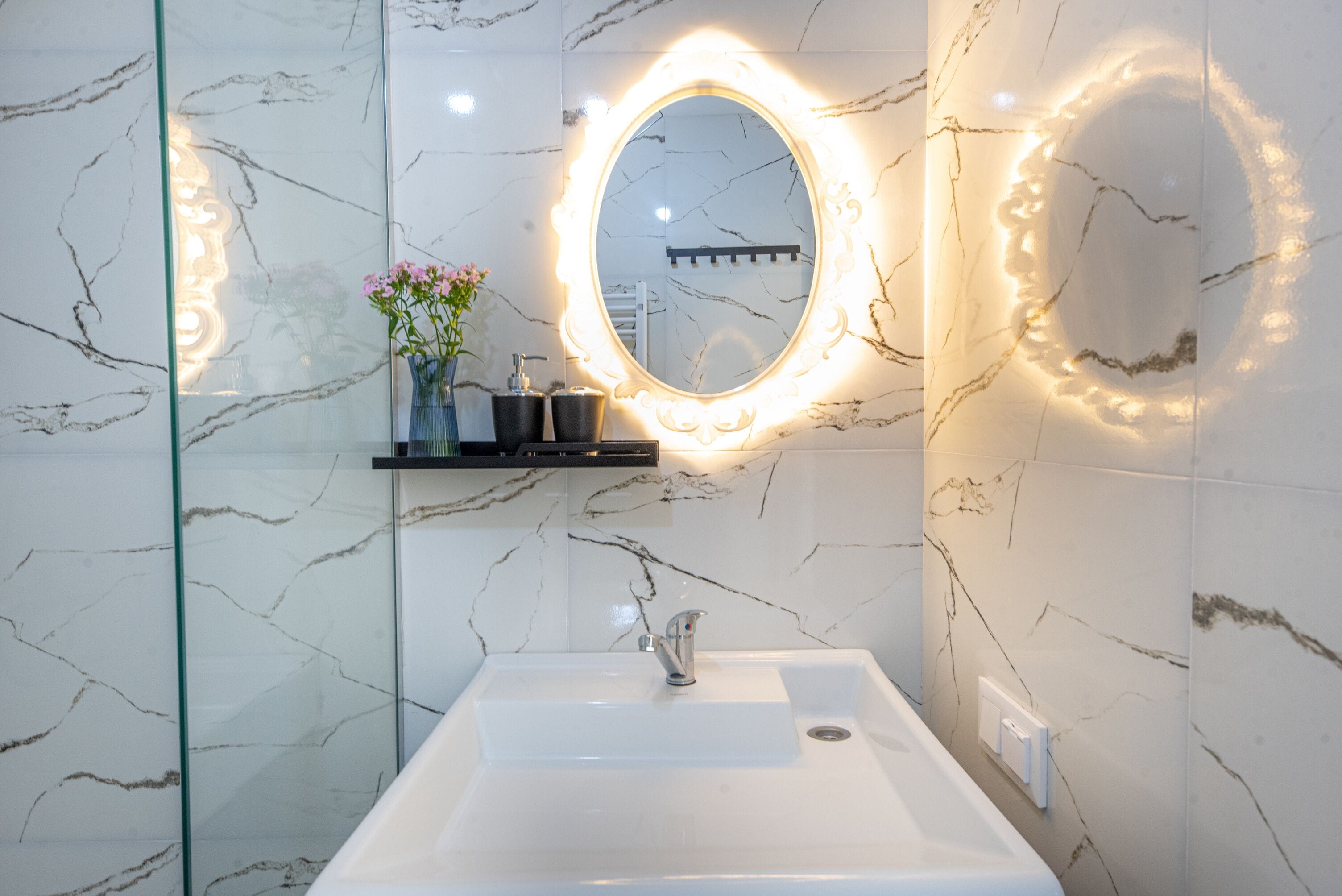 A bathroom with a white sink, a mirror with a light halo, and a vase with flowers.